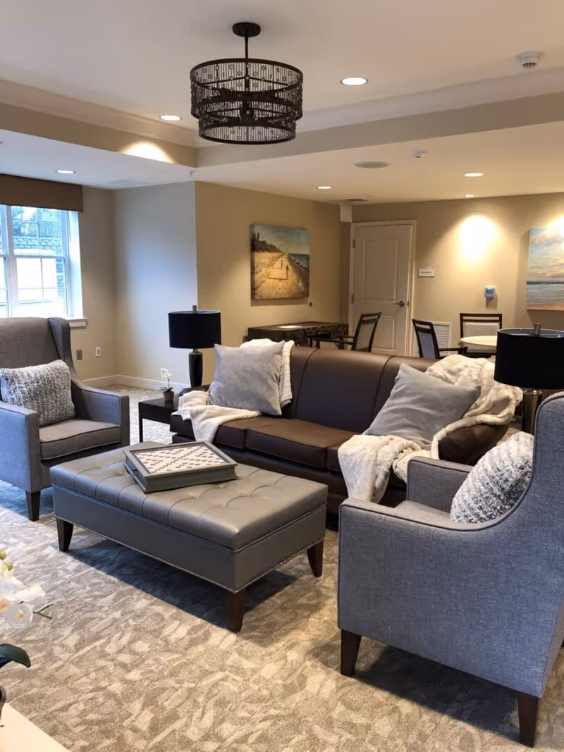 A cozy living room area with a brown leather sofa adorned with light-colored pillows and a throw blanket, flanked by two gray upholstered armchairs with patterned cushions. In front of the sofa is a tufted gray ottoman with a decorative tray on top. The room has beige walls with two landscape paintings, a window with a brown valance, and a modern black chandelier hanging from the ceiling. In the background, there is a small dining area with a round table and chairs.