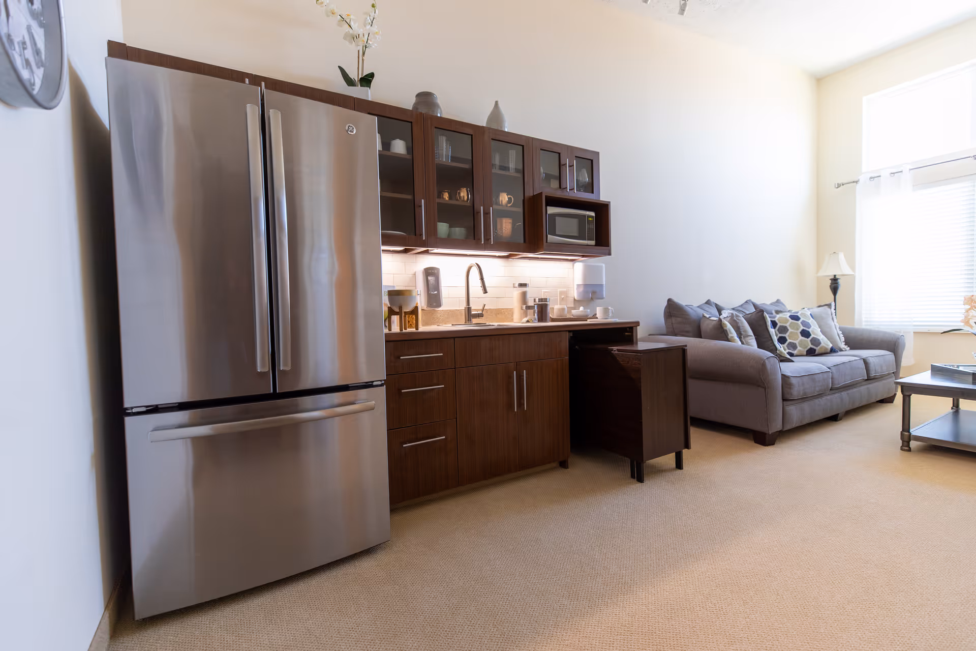 A modern kitchenette area with a stainless steel refrigerator, dark wood cabinets with glass doors, a microwave, and a sink. Adjacent to the kitchenette is a cozy living area with a gray sofa adorned with patterned pillows, a floor lamp, and a coffee table near a window with white blinds and curtains.
