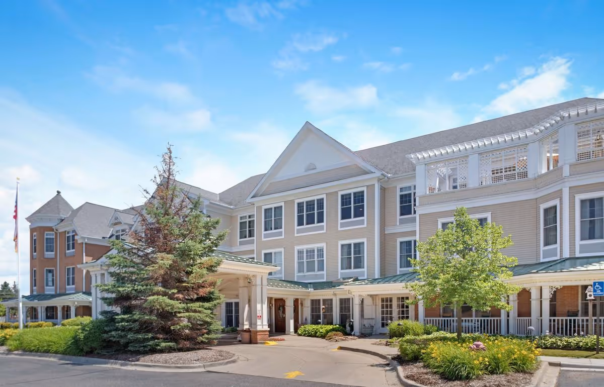Exterior view of a large senior living facility building with beige siding and white trim under a blue sky. The building has multiple windows, a covered entrance with columns, and landscaped greenery including trees and bushes around the driveway.