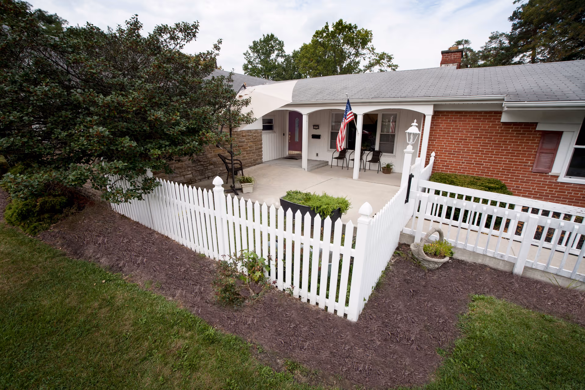 Front porch area of a single-story brick and stone house with a white picket fence surrounding a small garden. The porch has two chairs, an American flag, and a white lamp post near the fence. There are trees and shrubs around the house and a partly cloudy sky above.