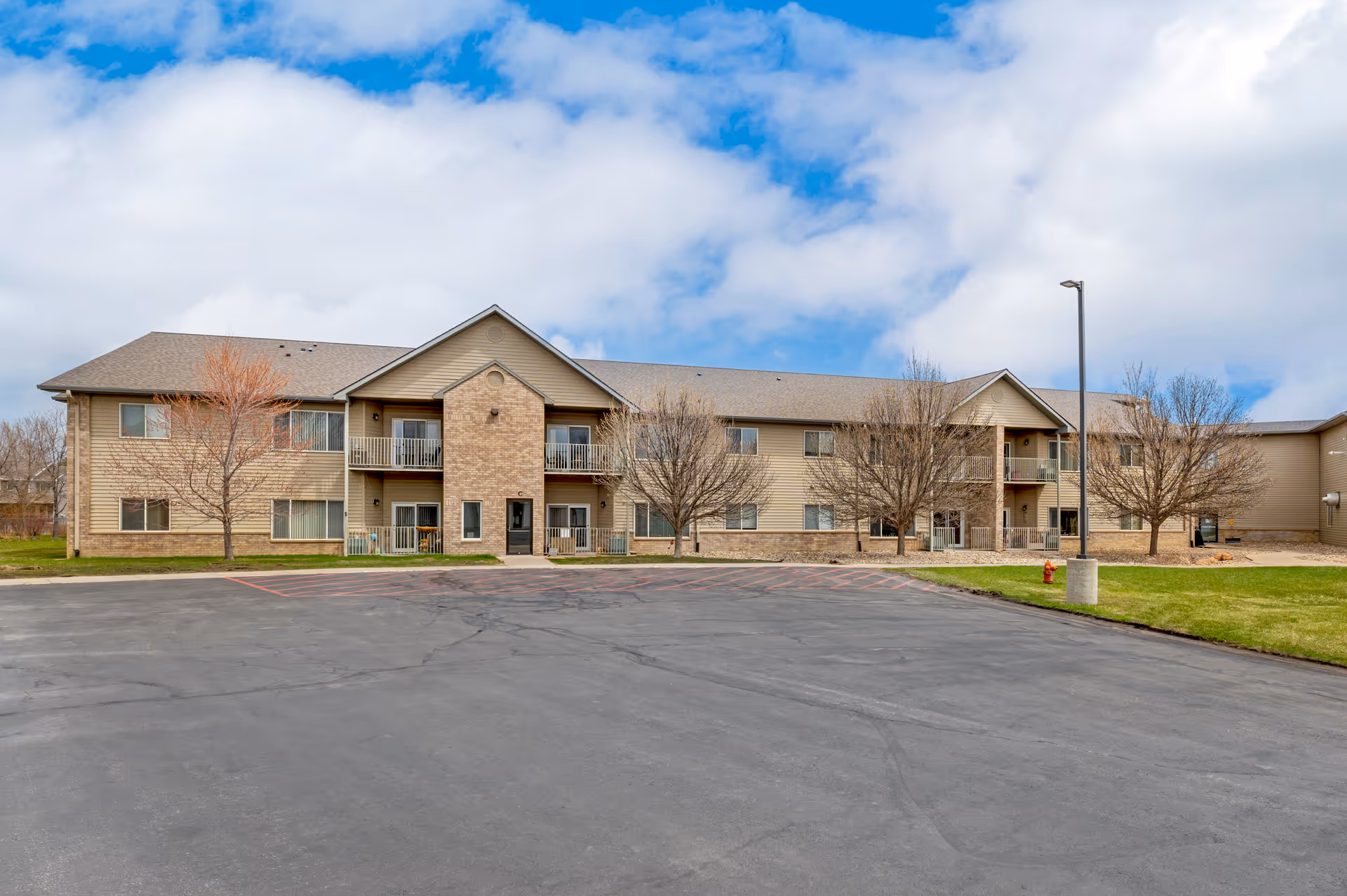 Exterior view of a two-story senior living facility building with beige siding and brick accents. The building has balconies, several windows, and a parking lot in front. Leafless trees and a grassy area surround the building under a partly cloudy sky.