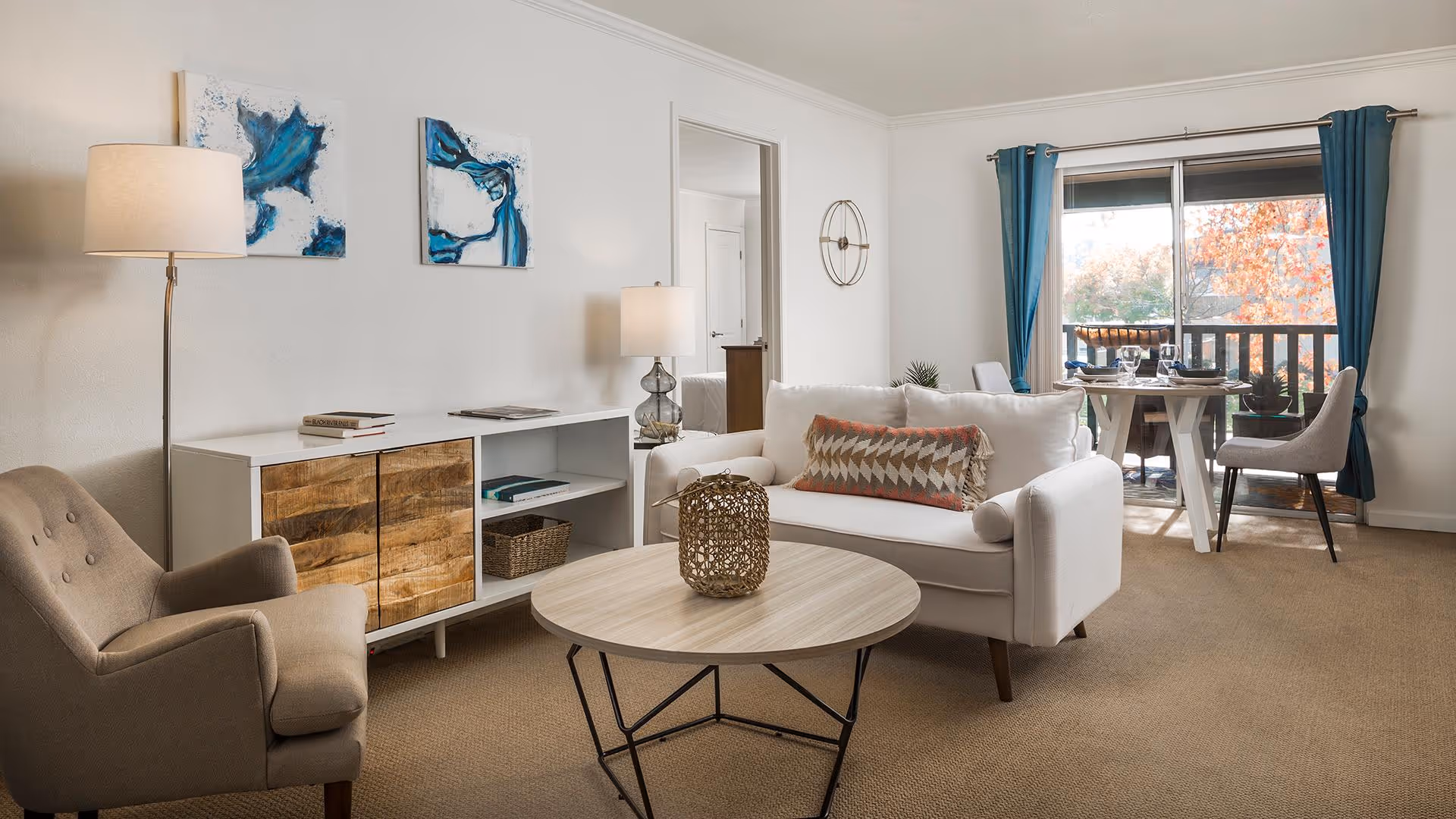 A bright living room with a white sofa adorned with a patterned pillow, a round wooden coffee table with a decorative item on top, a beige armchair, and a white sideboard with wooden doors. Two abstract blue and white paintings hang on the wall above the sideboard. A floor lamp and a table lamp provide lighting. In the background, there is a dining area with a round table set for four and large windows with blue curtains showing autumn foliage outside.