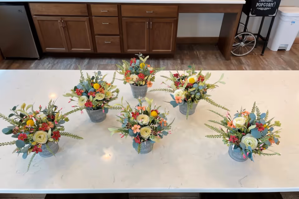 Five small flower arrangements with a mix of colorful flowers and greenery placed on a white marble countertop in a kitchen area with wooden cabinets and a popcorn machine in the background.