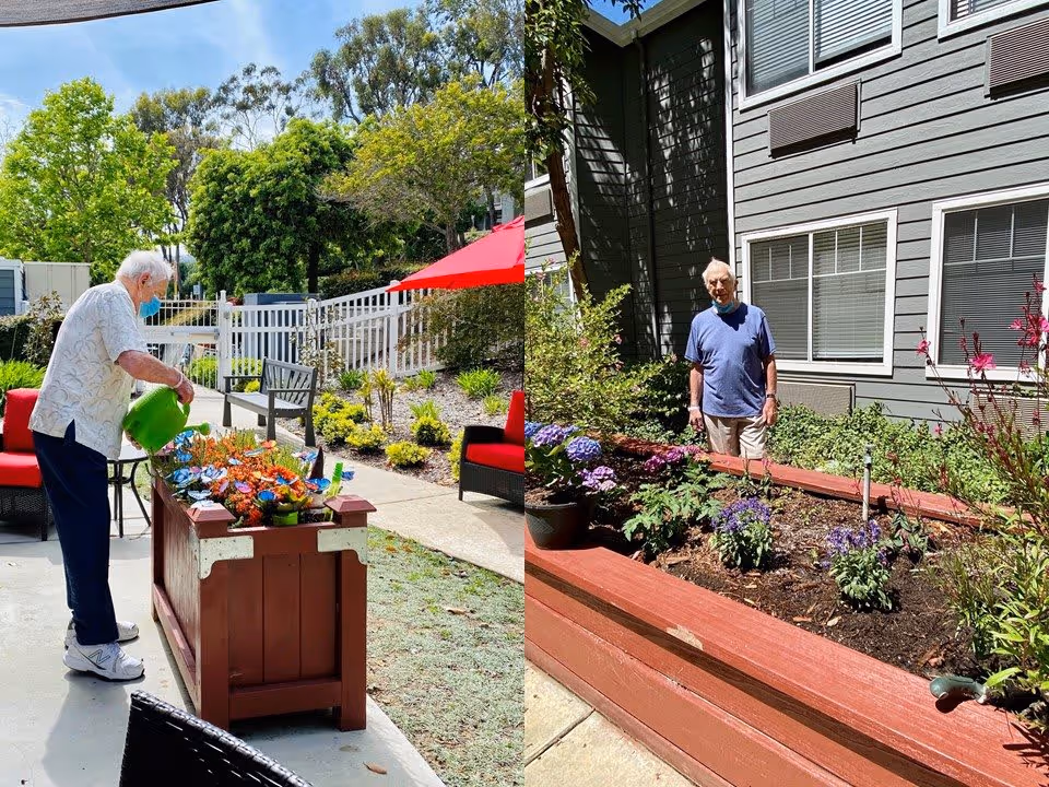 Two elderly men tending to garden beds in an outdoor patio area of a senior living facility. One man is watering plants with a green watering can, while the other stands among various flowering plants. The area is surrounded by greenery, a white fence, and outdoor seating with red cushions.