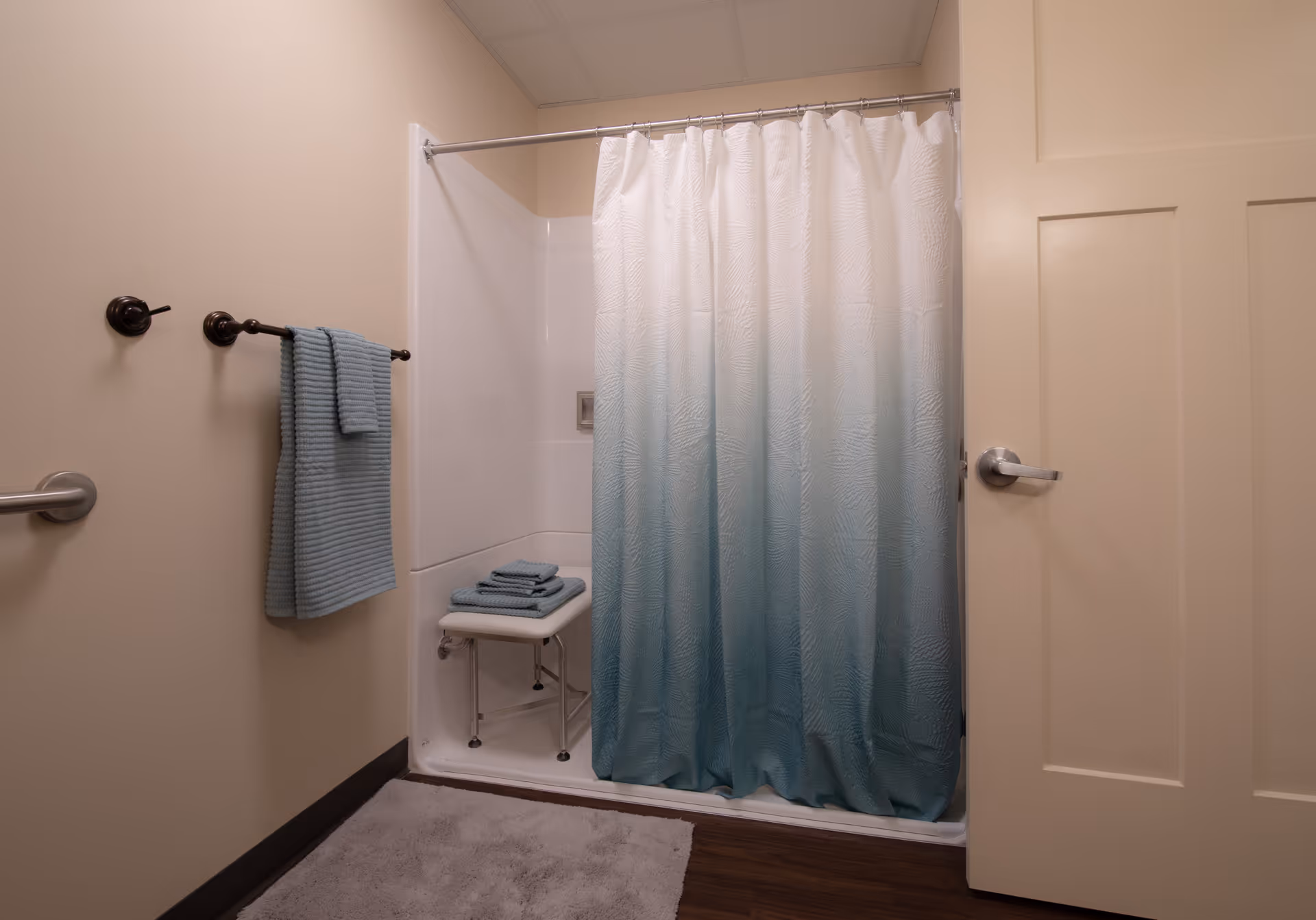 A bathroom shower stall with a teal ombre curtain, a shower chair with folded towels, towel rack, grab bar, and an open door.