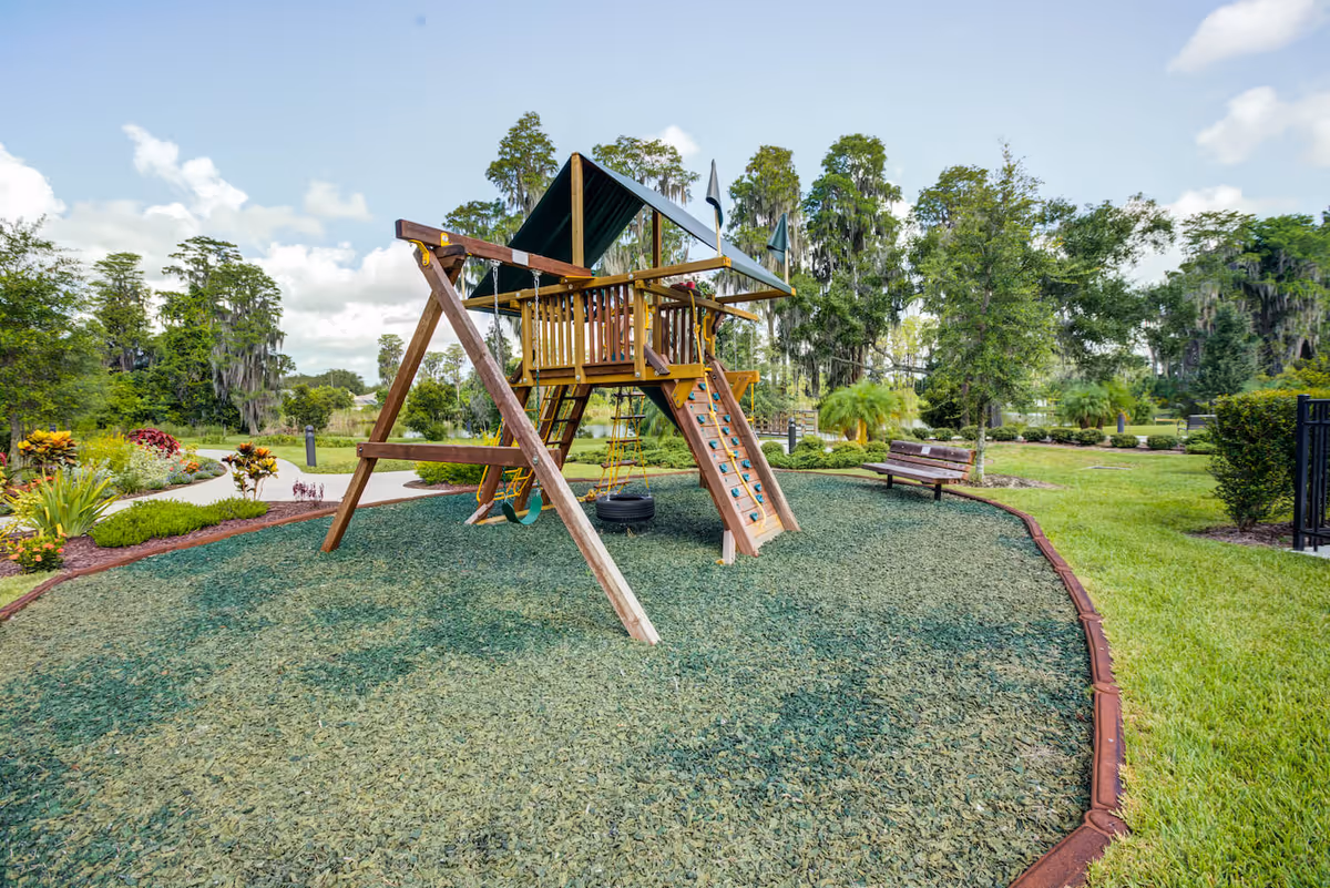 Outdoor playground area with a wooden playset featuring swings, a climbing wall, and a covered platform, surrounded by green landscaping and trees under a partly cloudy sky.