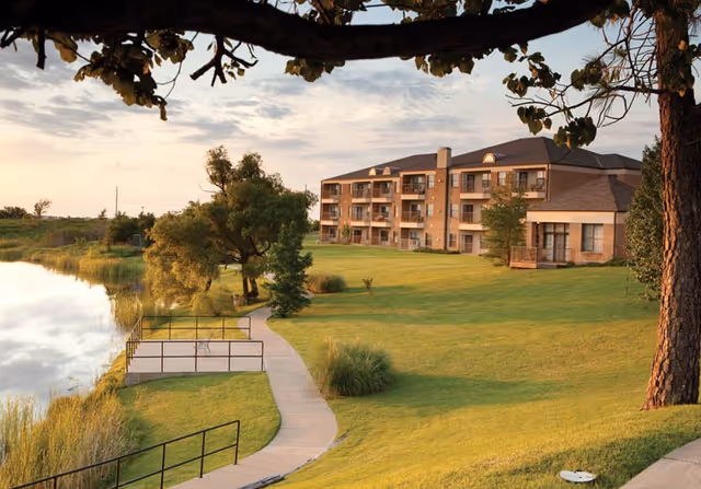 A large senior living facility building with multiple balconies overlooking a grassy area and a pond. A paved walkway curves along the edge of the pond, surrounded by trees and greenery under a partly cloudy sky.