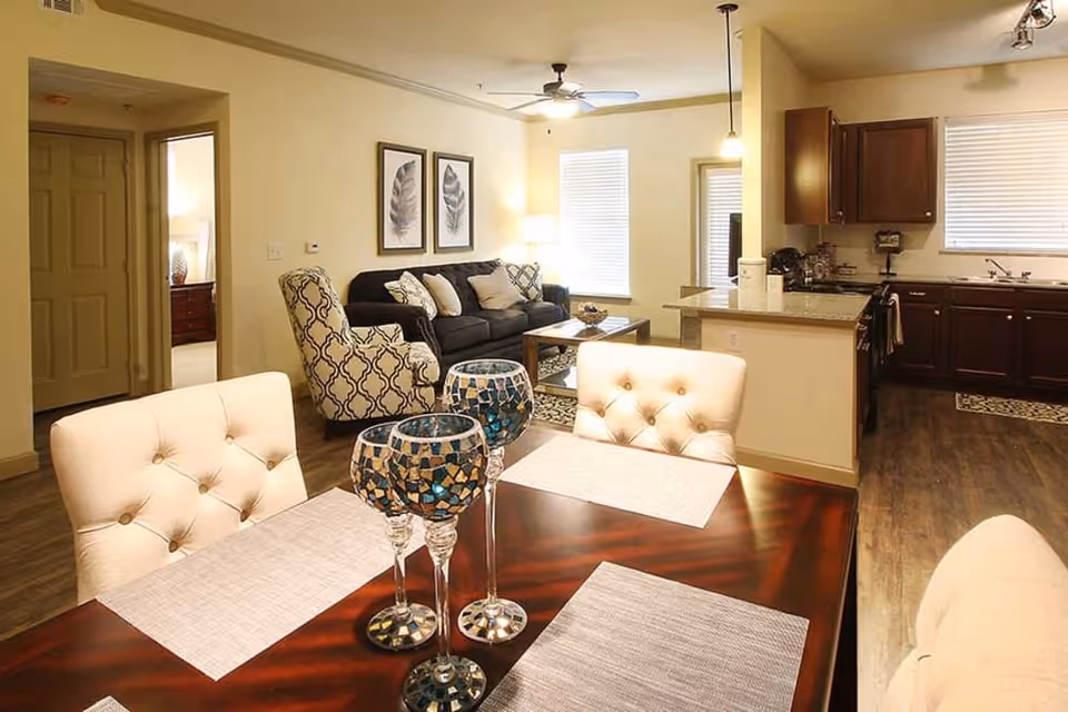 Open-plan dining and living area featuring a dark wood dining table with tufted chairs and decorative mosaic candle holders, a sofa and patterned armchair, and a kitchen with dark cabinets in the background.