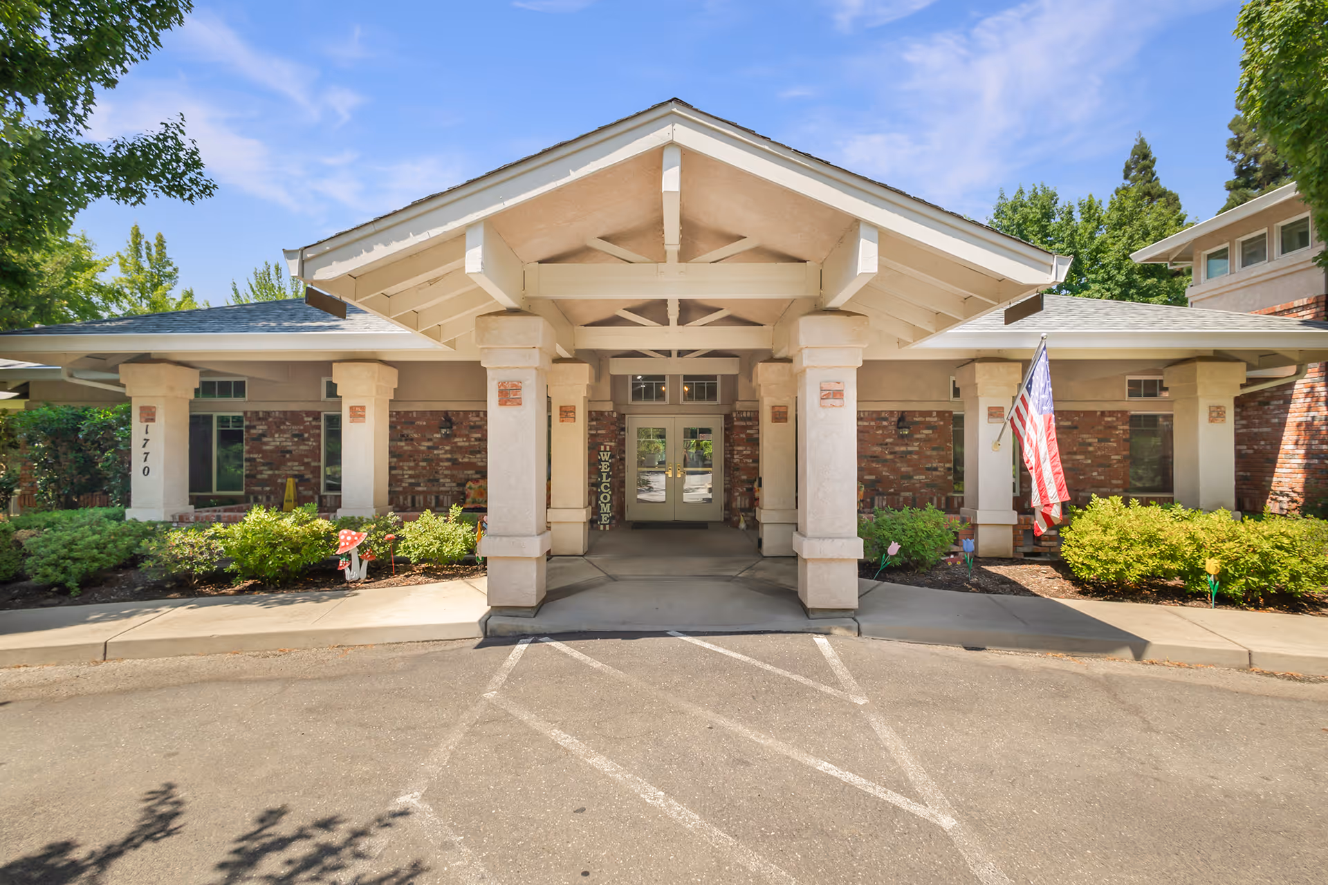 Front entrance of a senior living facility with a covered porte-cochère, columns, landscaping, and an American flag.