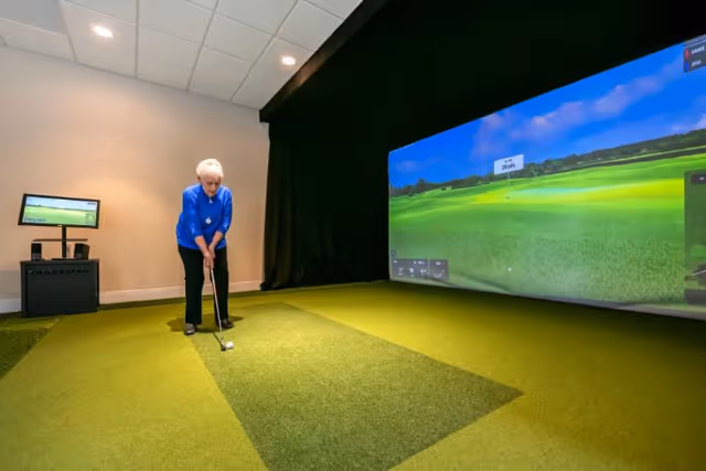 An elderly woman playing indoor golf using a golf simulator in a room with green carpet resembling a putting green and a large screen displaying a golf course.