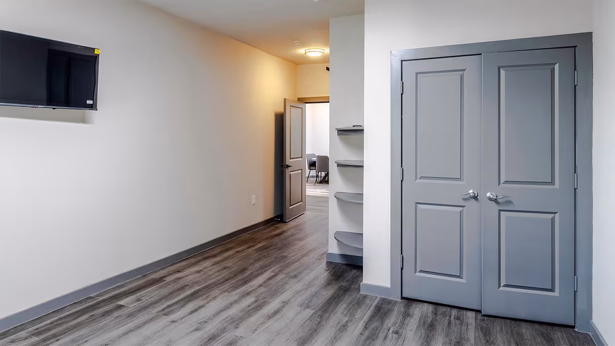 Interior hallway with gray double doors on the right, a wall-mounted TV on the left, and an open door at the end leading to a room with chairs and a table.