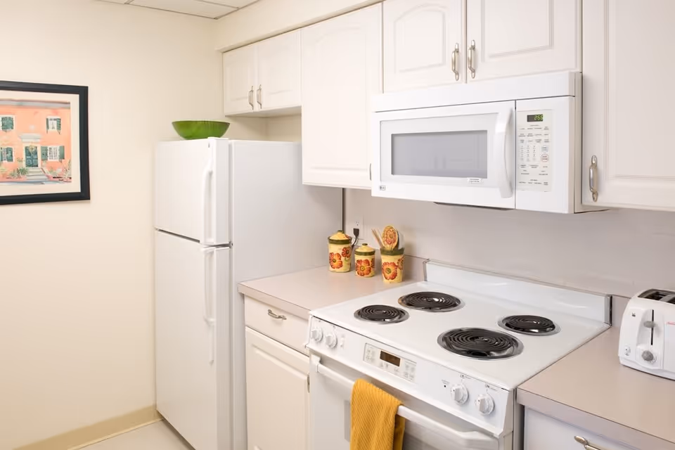 A clean kitchen with white cabinets, a white refrigerator, a white microwave mounted above a white electric stove with four coil burners. There is a yellow towel hanging on the oven handle, three decorative canisters with floral designs on the countertop, a white toaster, and a framed picture on the wall.
