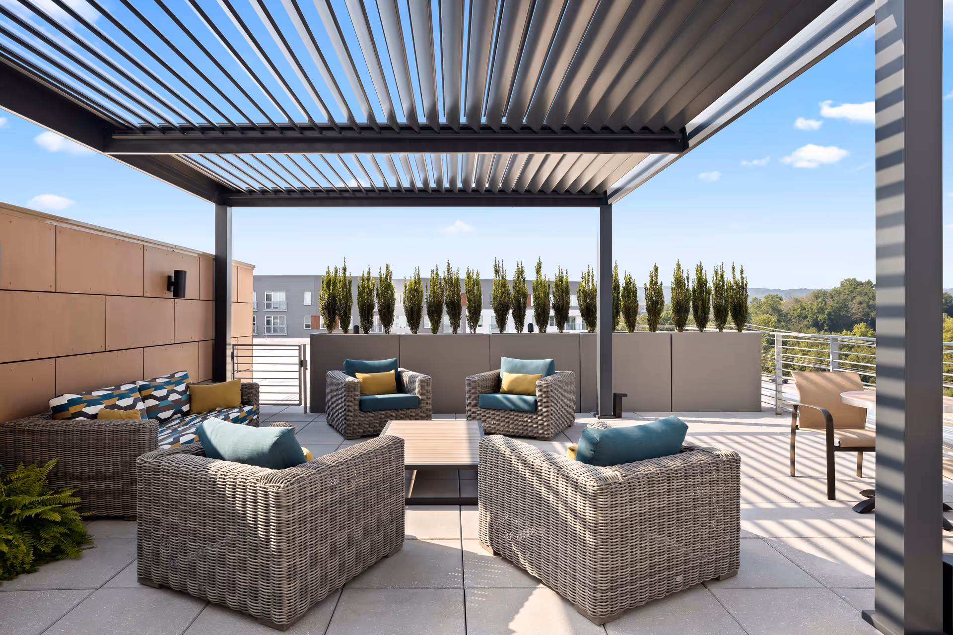 Outdoor patio area with modern wicker furniture including a sofa and four armchairs with blue and yellow cushions arranged around a low rectangular coffee table. The space is shaded by a slatted pergola roof and has tall potted plants along the railing. The background shows a clear sky and distant trees.