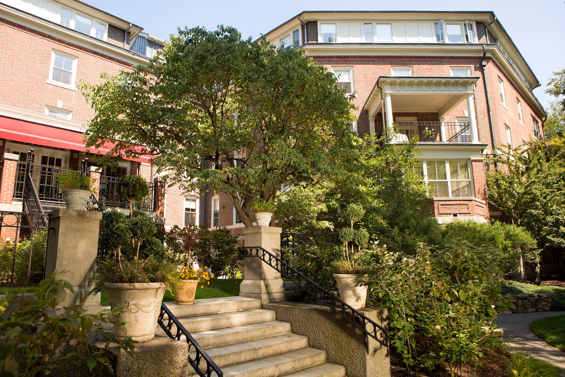 Exterior view of a multi-story brick building with a red awning, surrounded by lush green trees and plants. Stone steps with black iron railings lead up to the entrance, which is partially visible. The scene is bright and sunny.