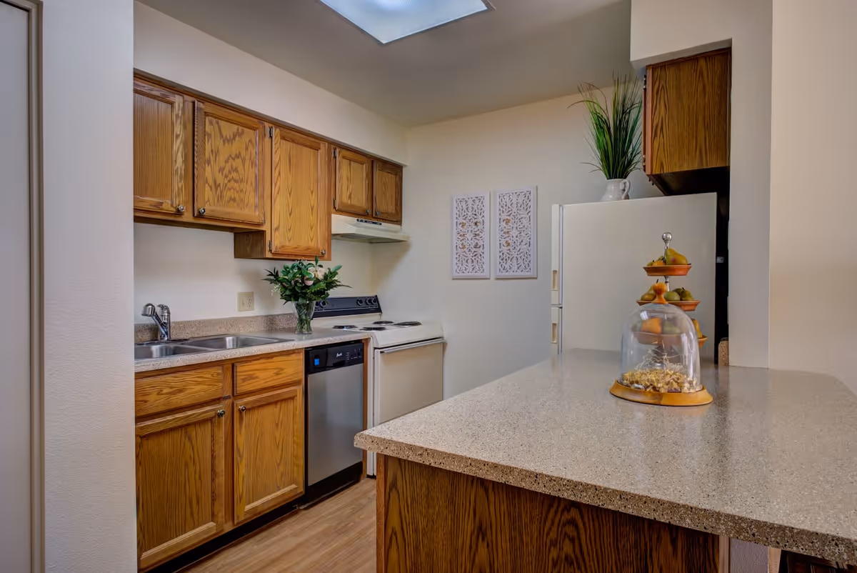 A kitchen with wooden cabinets, a double sink, a dishwasher, an electric stove with a range hood, a refrigerator, and a countertop with a tiered fruit stand and a glass dome containing snacks. There is a plant on the refrigerator and two decorative wall hangings on the wall.