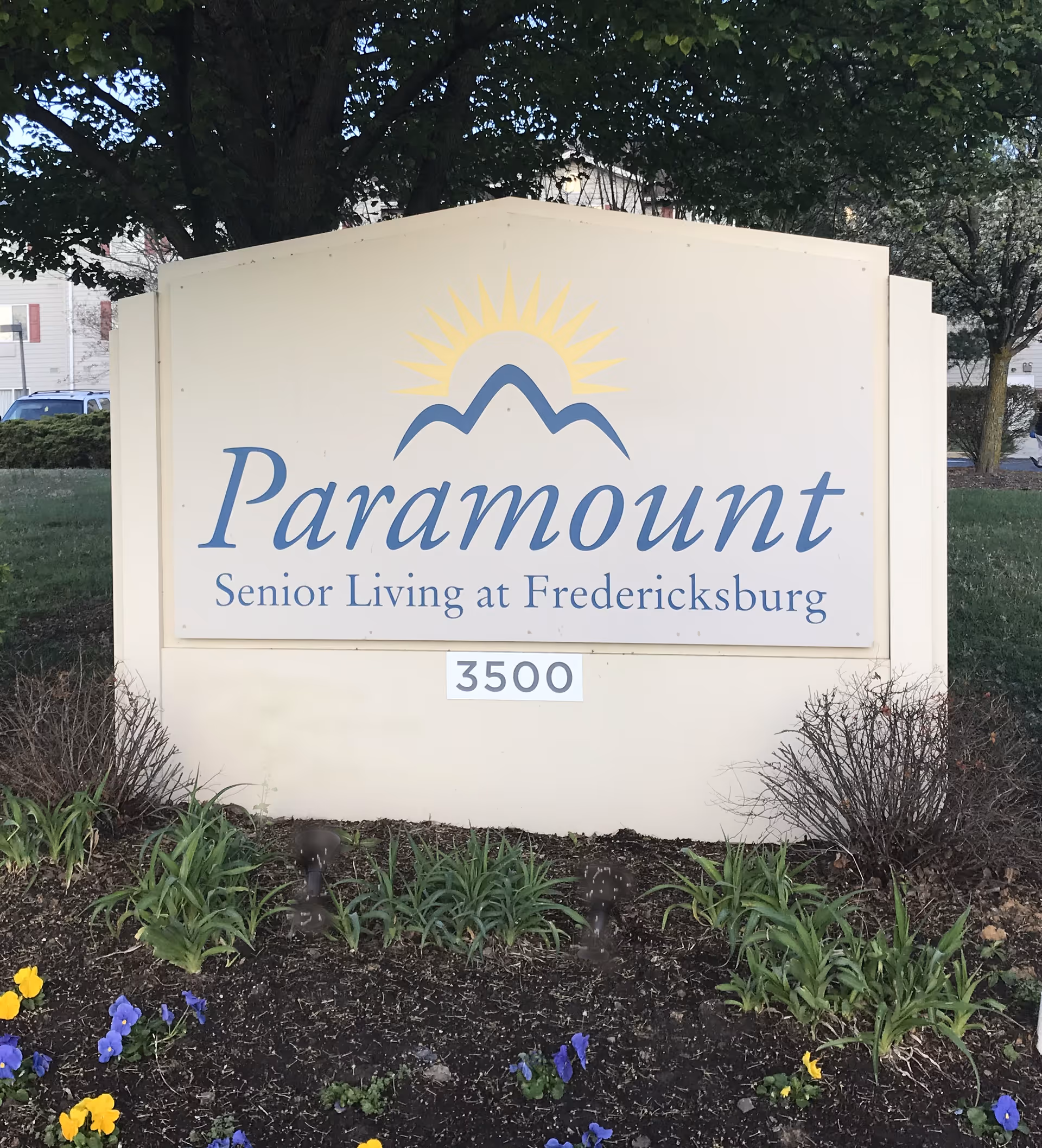 Outdoor sign for Paramount Senior Living at Fredericksburg with the address number 3500, surrounded by plants and flowers, with trees and buildings in the background.