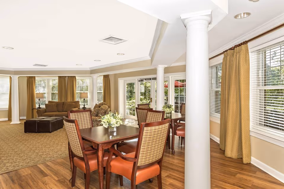 Bright communal dining and sitting area with a wooden table and chairs, sofas, large windows, and decorative columns.