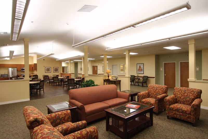 Furnished communal lounge with sofas and armchairs around a coffee table, with a dining area and kitchenette visible in the background.