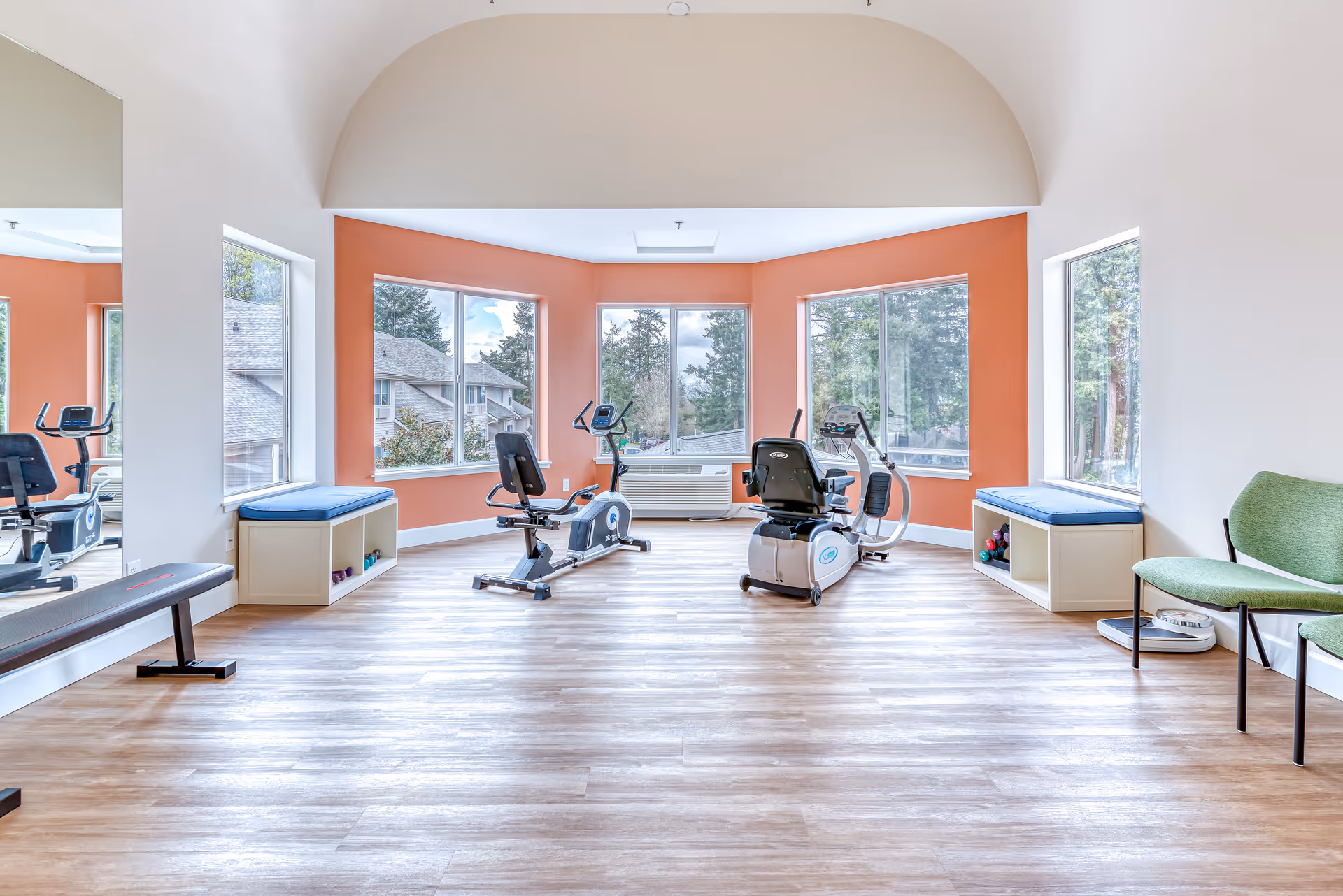Bright exercise room with large windows showing outdoor trees and buildings. The room has light wood flooring, orange accent walls, and white ceiling with an arch. There are two recumbent exercise bikes in the center, two cushioned benches with storage underneath on either side, a green chair, a scale, and a large mirror on the left wall.