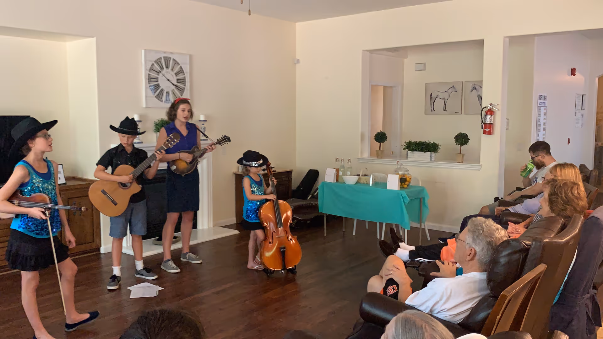 A group of four children performing music with instruments including a violin, guitar, mandolin, and cello in a living room area. Several elderly people are seated in chairs watching the performance. The room has wooden floors, light-colored walls, and a table with refreshments covered by a green tablecloth in the background.