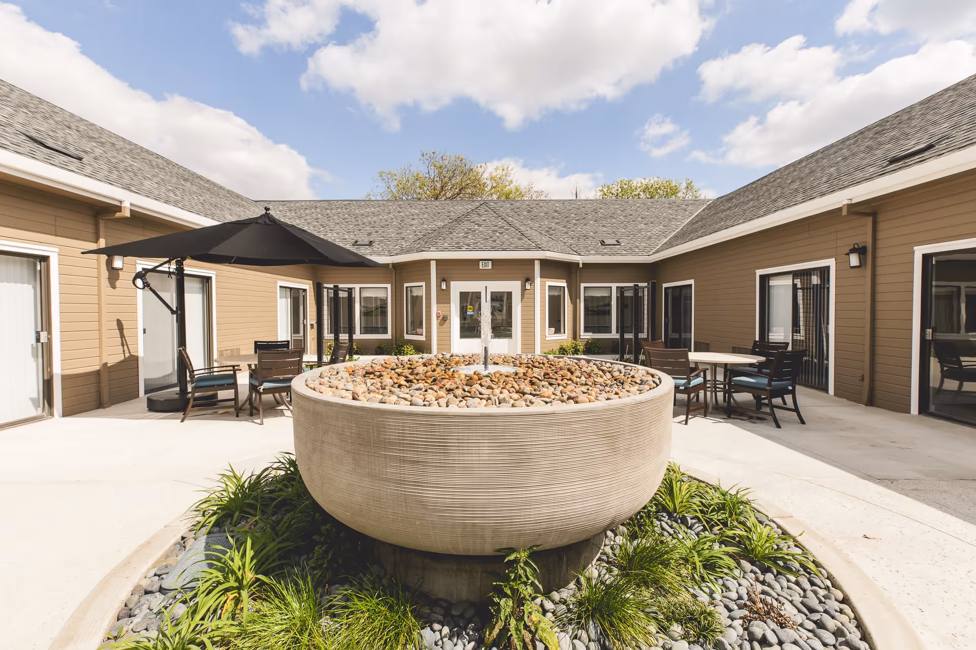 Outdoor courtyard area of a senior living facility with a large round stone water fountain filled with pebbles in the center, surrounded by green plants and a circular stone border. The courtyard is enclosed by a single-story building with beige siding and multiple sliding glass doors. There are several tables and chairs with umbrellas for shade placed around the courtyard. The sky is partly cloudy.