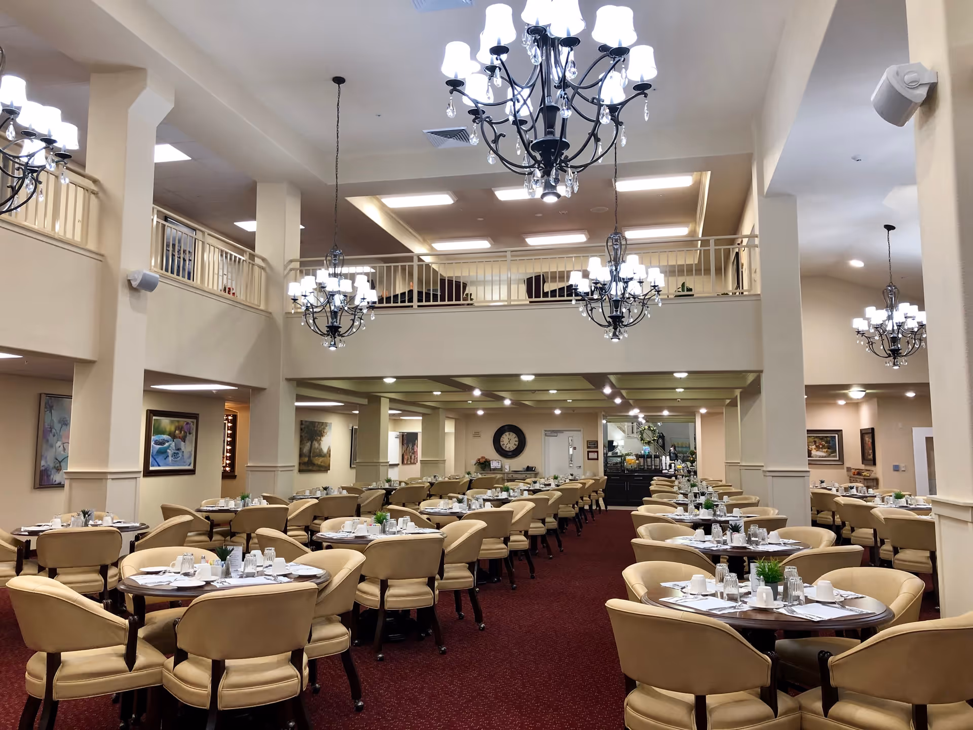 Spacious dining room with multiple round tables set with white cups, glasses, and silverware. Beige cushioned chairs surround each table. Elegant chandeliers hang from the ceiling, and there is a second-floor balcony overlooking the dining area. The room has beige walls and a red carpeted floor.