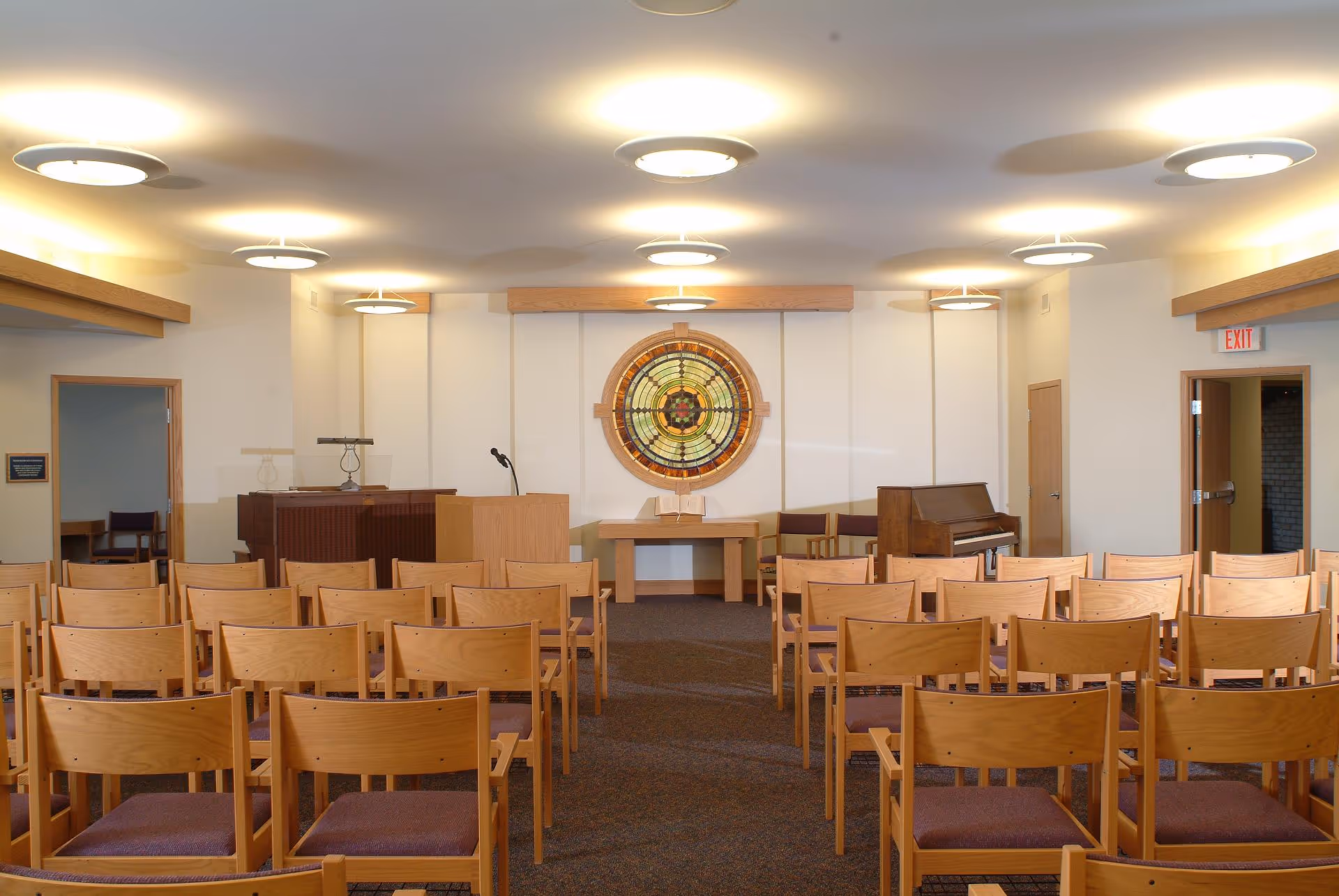 Interior view of a chapel or meeting room with rows of wooden chairs facing a podium and a table with an open book. A stained glass circular window is centered on the back wall above the table. There is a piano to the right and a lectern to the left. The room is well-lit with ceiling lights and has doors on both sides.