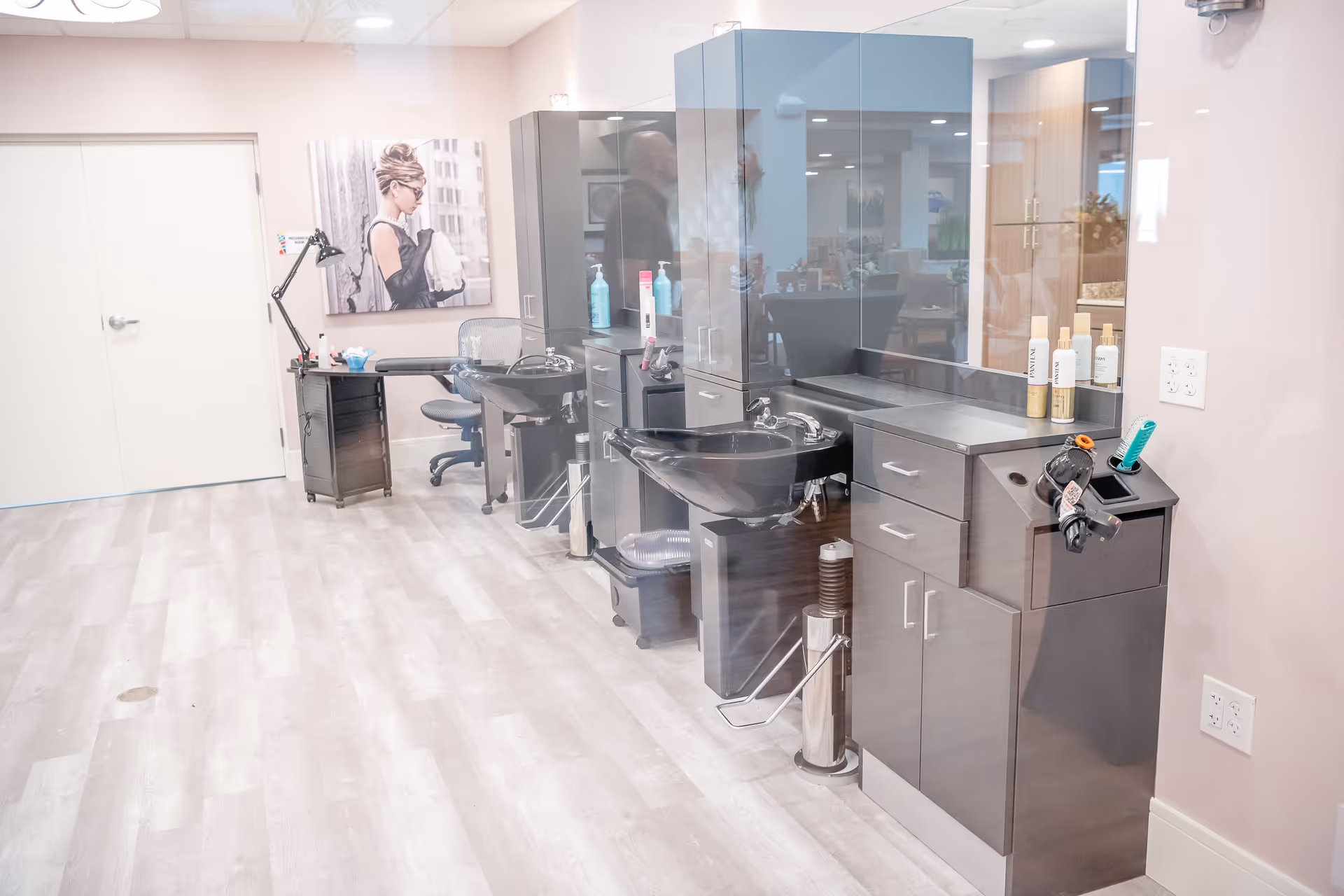 Interior view of a modern hair salon area within a facility, featuring black shampoo sinks with chairs, dark cabinetry, hair care products on the counter, a desk with a lamp and chair, and a large wall photo of a woman with an updo hairstyle.