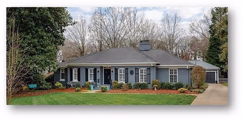 Single-story residential building with a gray roof and blue-gray exterior walls, surrounded by trees and a well-maintained lawn with shrubs and plants. A driveway leads to a garage on the right side of the house.