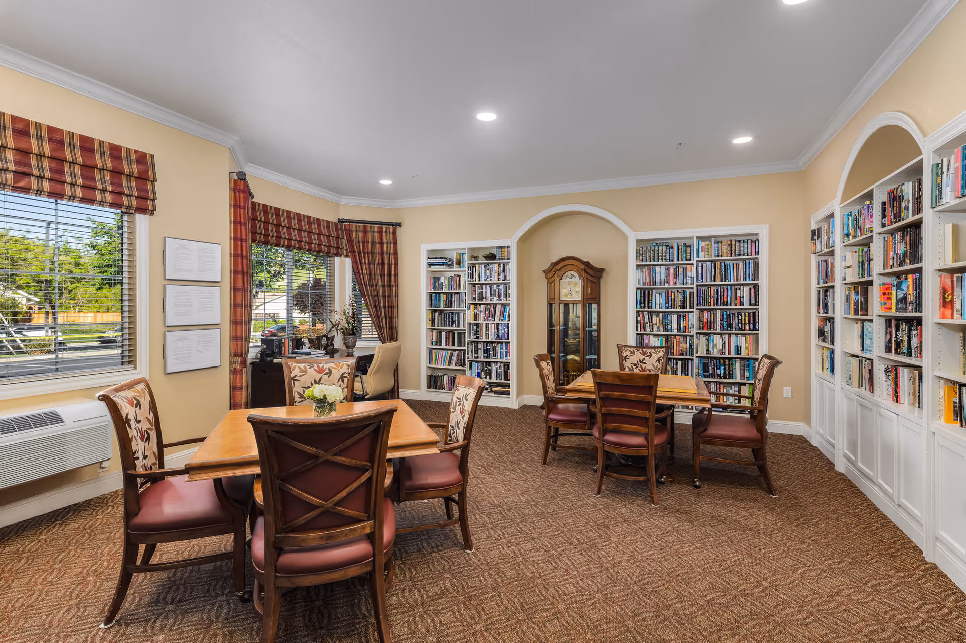 A cozy library room in a senior living facility with two wooden tables surrounded by chairs with floral upholstery. The walls are lined with built-in bookshelves filled with books, and a grandfather clock is placed in a recessed archway. The room has large windows with plaid curtains and valances, beige walls, and patterned carpet flooring.