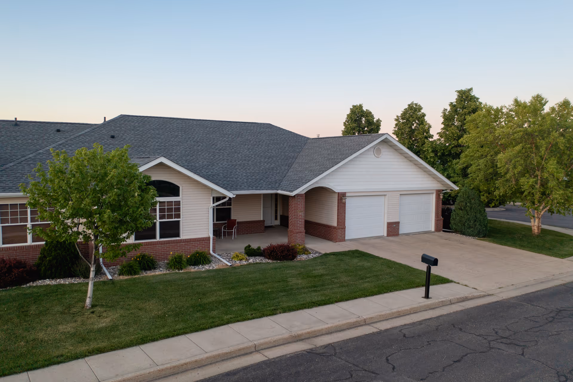 Exterior view of a single-story residential building with a two-car garage, a front porch with chairs, a well-maintained lawn, and trees along the sidewalk and driveway.