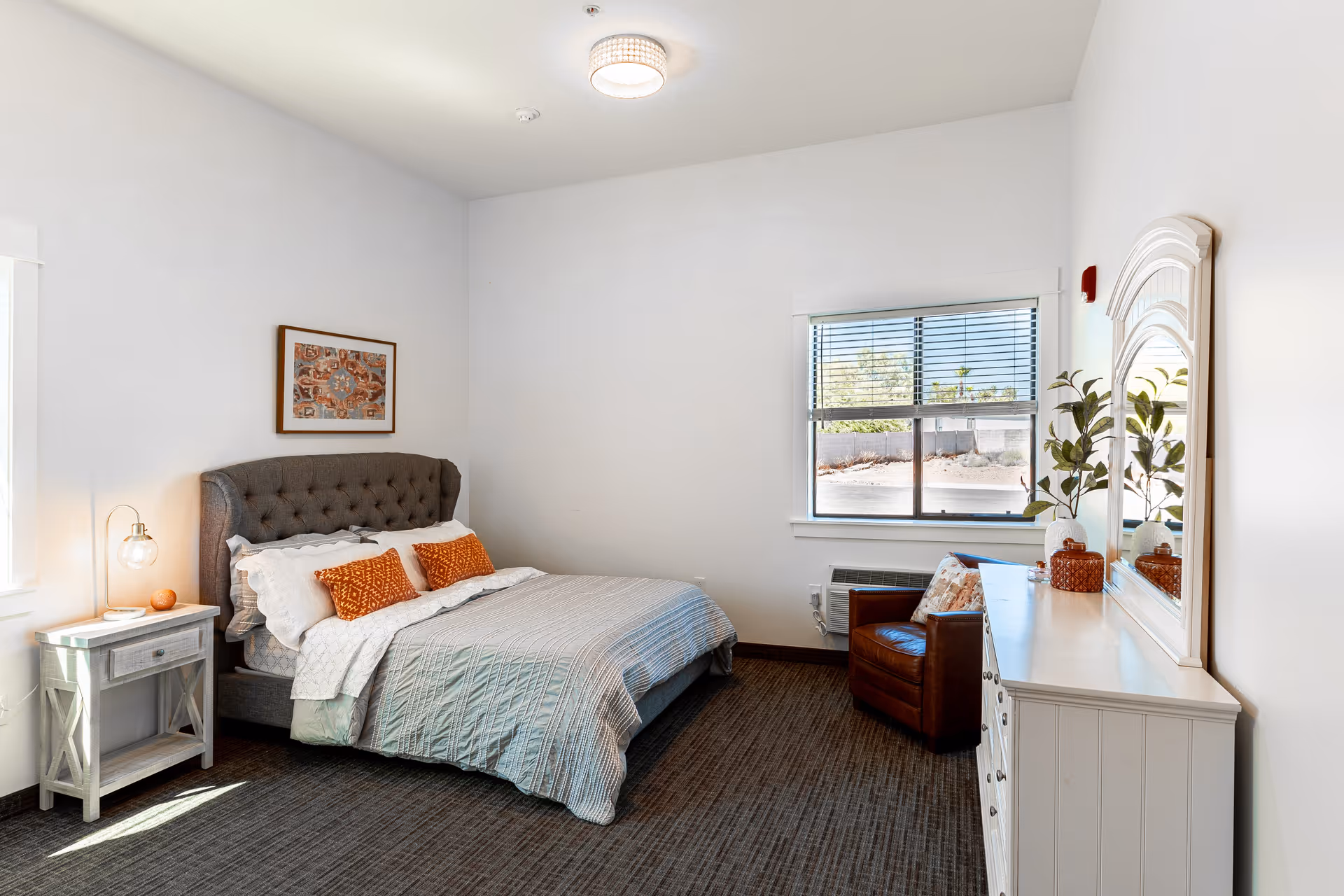 Bright furnished bedroom with a tufted headboard bed, nightstand with lamp, dresser and mirror, and a window.