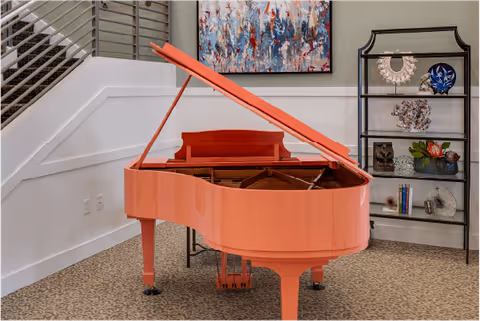 A coral-colored grand piano with its lid open is placed on a patterned carpet in a room. Behind the piano, there is a modern metal staircase railing on the left and a decorative black metal shelving unit on the right holding various art pieces and books. A colorful abstract painting hangs on the wall above the piano.