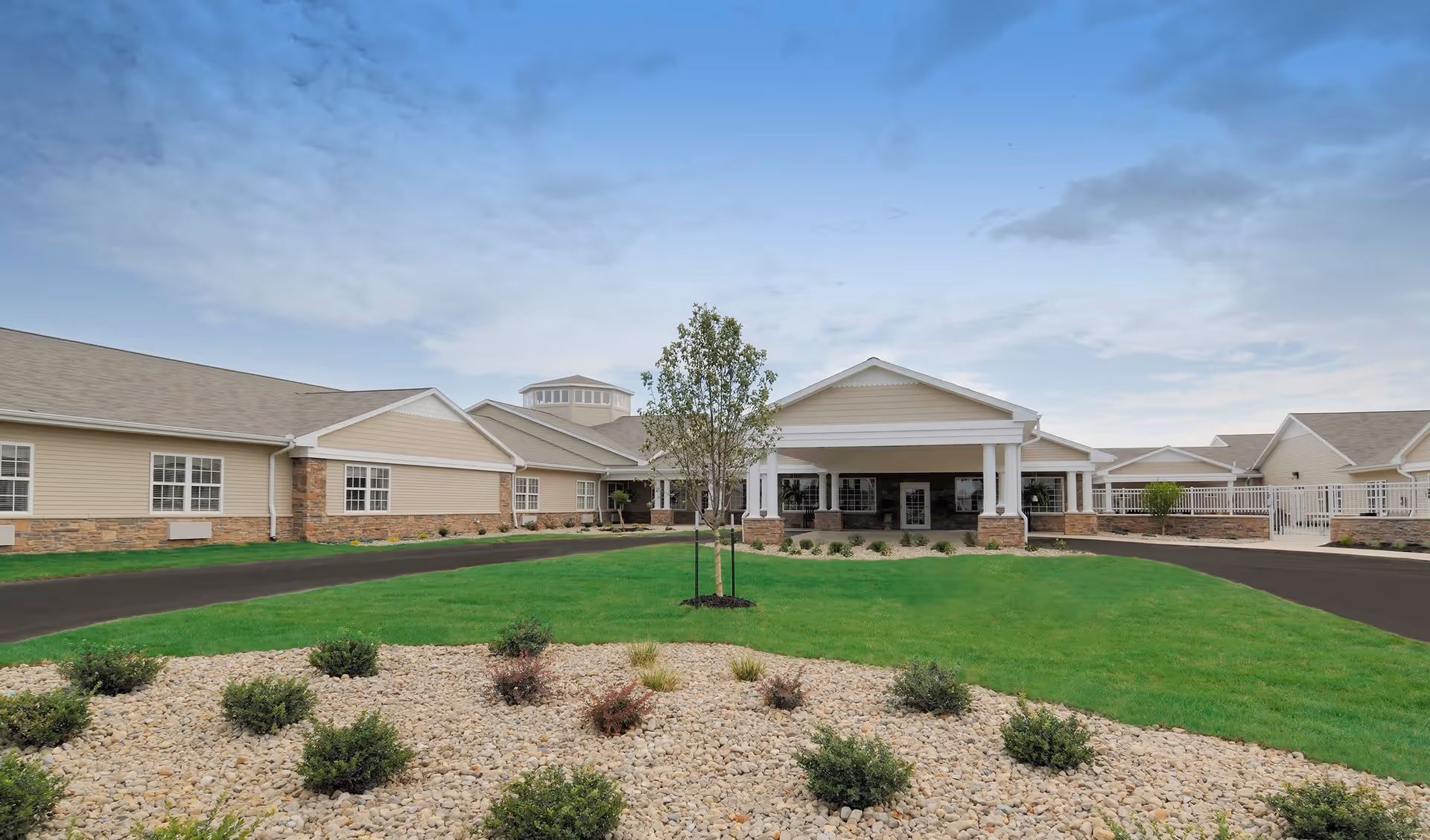 Exterior view of Orchard Grove Senior Living facility showing a single-story building with beige siding, stone accents, a covered entrance with white columns, a paved driveway, green lawn, and landscaped area with small bushes and rocks under a partly cloudy sky.
