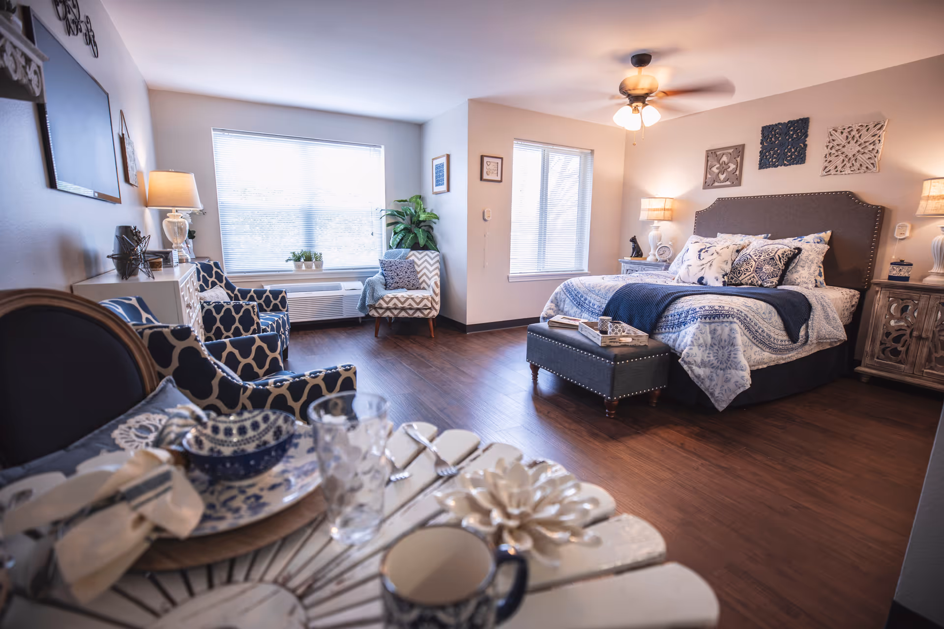 A cozy, well-lit bedroom with a large bed adorned with blue and white patterned bedding and multiple pillows. The room features wooden flooring, two windows with blinds, a ceiling fan with lights, and decorative wall art above the bed. There are two bedside tables with lamps and decorative items, a cushioned bench at the foot of the bed, and several patterned armchairs near the windows. A small dining table with place settings is visible in the foreground.