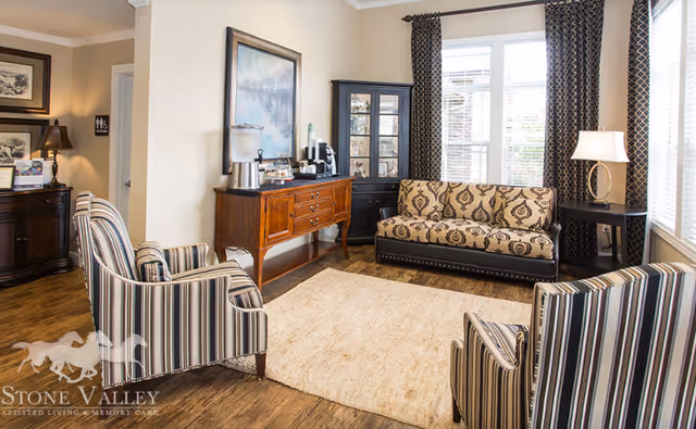 A cozy living room area with a patterned sofa, two striped armchairs, a wooden sideboard with a coffee and tea setup, a black display cabinet, a side table with a lamp, and large windows with patterned curtains letting in natural light. The floor is wooden with a beige area rug in the center.
