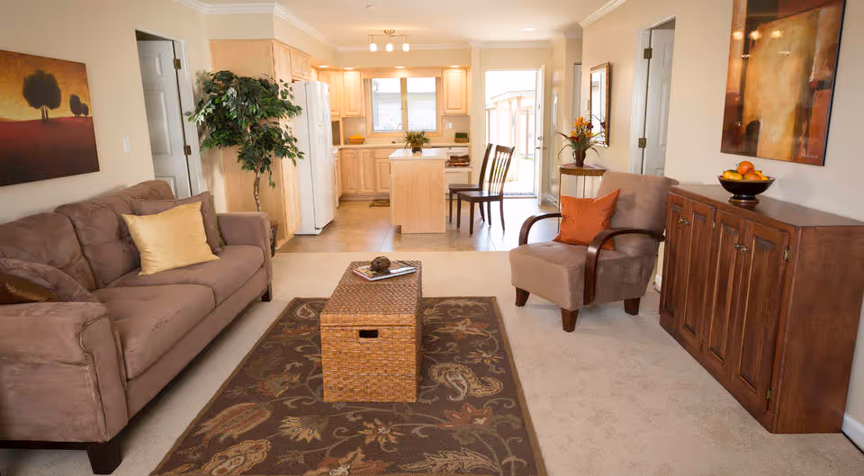 Bright open living room with a sofa, armchair, wicker coffee table and a view into a light-wood kitchen.