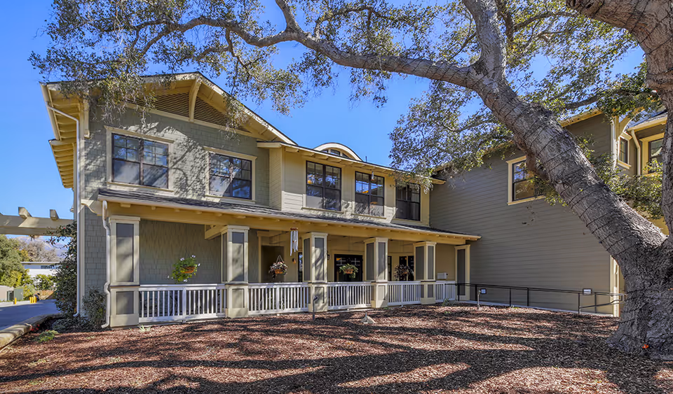 Exterior view of a two-story building with green and beige siding, multiple windows, a covered porch with white railing, hanging flower baskets, and a large tree casting shadows on the ground in front of the building under a clear blue sky.