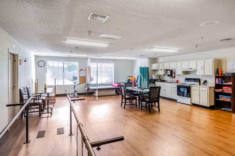 A spacious room with wooden flooring featuring a kitchen area with white cabinets, stove, and sink on the right side. In the center, there is a round table with four chairs. On the left side, there are parallel bars for walking exercises, several chairs, and a hospital bed near large windows with blinds. The room is well-lit with ceiling lights and has a clock on the wall.