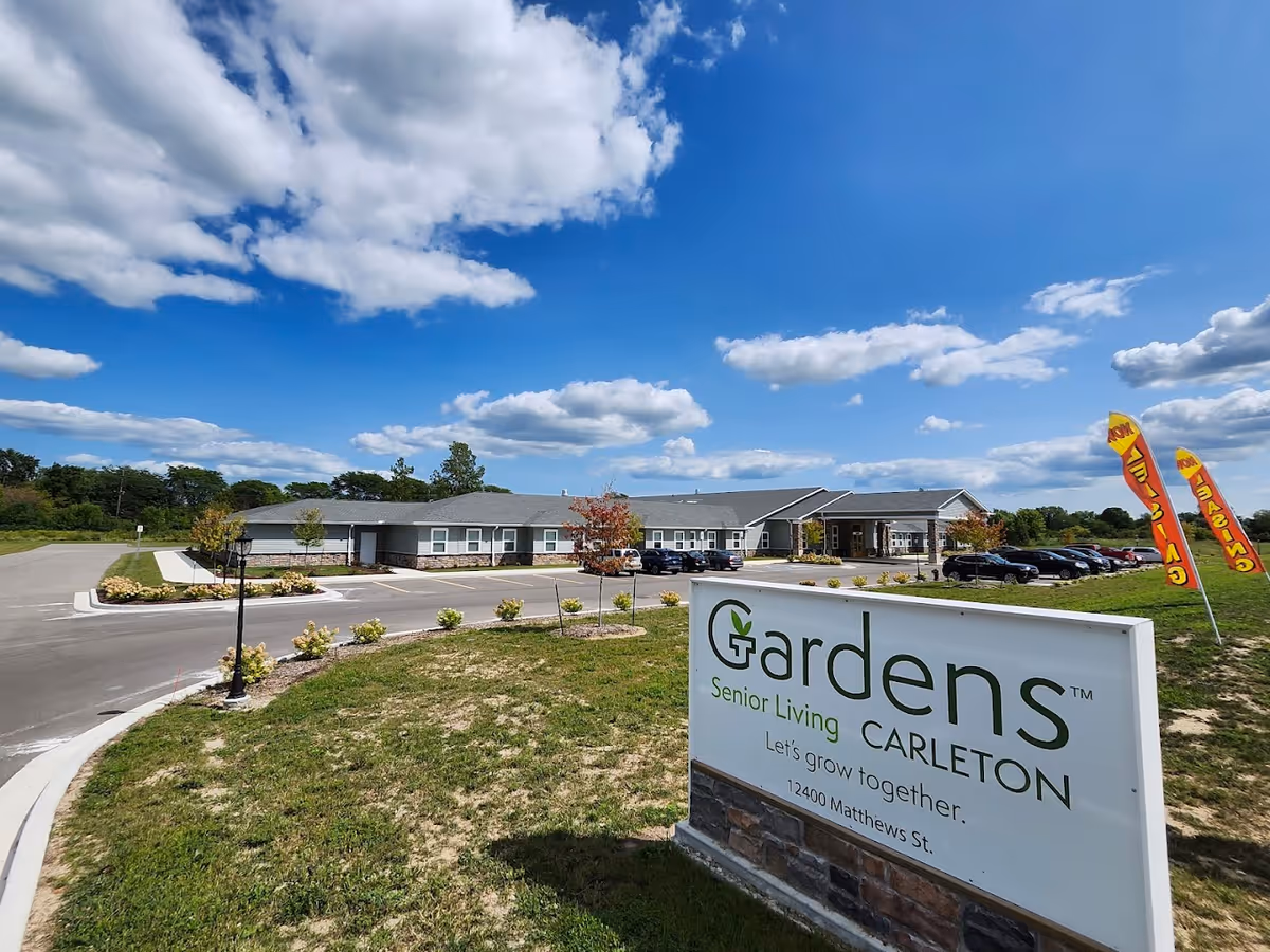 Exterior view of the Gardens Senior Living Carleton building with its sign in the foreground, parking lot and entrance under a blue sky.