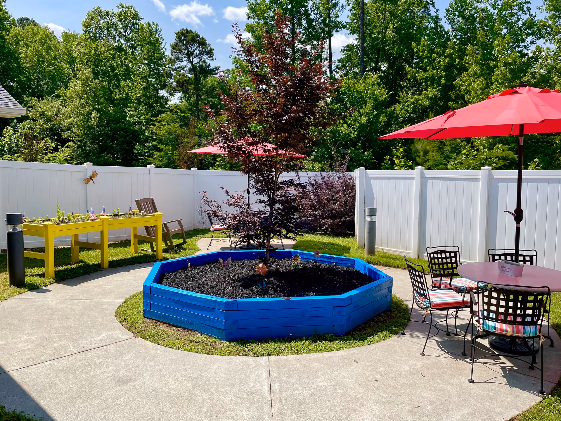 Outdoor garden area at Franklin Manor Assisted Living Center featuring a blue octagonal raised flower bed with a small tree in the center, a yellow raised planter box with small plants and American flags, a wooden rocking chair, and a patio table with four chairs under a red umbrella. The area is surrounded by a white fence and lush green trees under a partly cloudy sky.