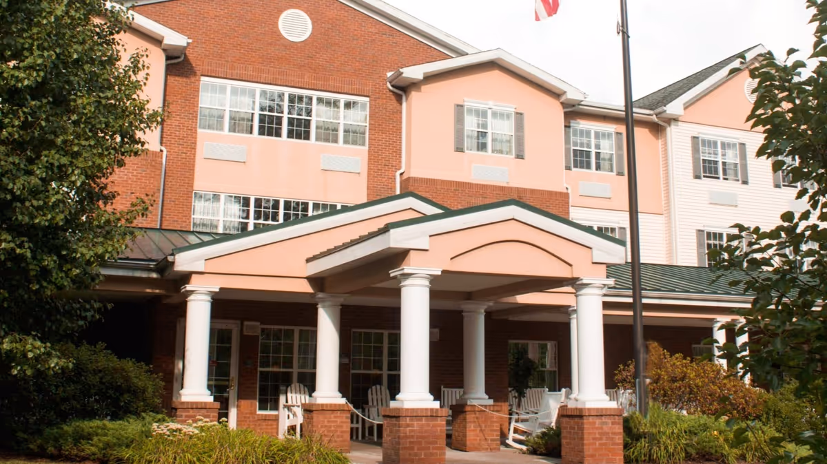 Front entrance of a multi-story senior living building with a covered portico supported by white columns, brick and pink facade, and surrounding landscaping.