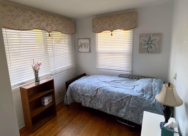 Small bedroom with a single bed covered in a blue patterned comforter, two windows with blinds and valances, a small bookshelf and a bedside lamp.