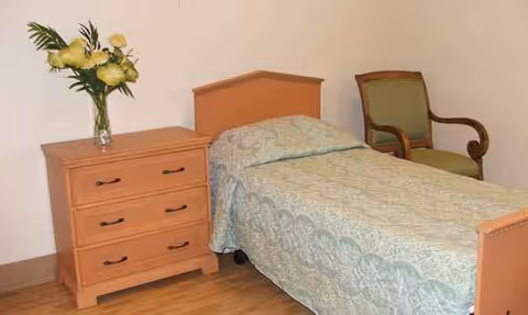 A bedroom with a single bed, dresser with a vase of flowers, and a green chair.