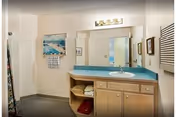 A clean bathroom with a large mirror above a wooden vanity with a blue countertop and a white sink. The vanity has open shelves with folded towels. There is a towel rack on the wall with a towel hanging, and a framed picture above it. The floor is tiled in a dark color, and the walls are light-colored. A shower area is partially visible on the left side.