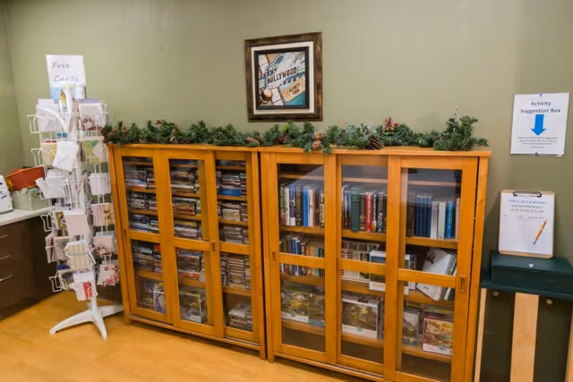 A room corner with a wooden cabinet containing books and DVDs behind glass doors. On top of the cabinet is a decorative garland with pine cones. Above the cabinet hangs a framed Hollywood-themed picture. To the left is a white rotating rack with cards labeled 'Free Cards'. To the right is an activity suggestion box with a sign above it on the wall.