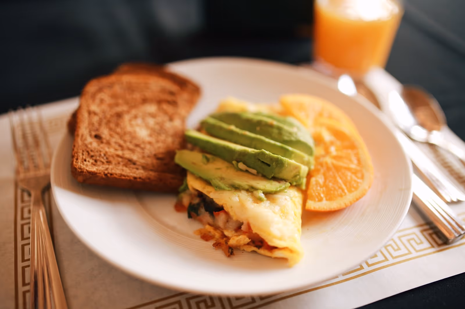 A plate with a slice of omelette topped with sliced avocado, two slices of toasted bread, and two orange slices. A glass of orange juice and silverware are placed beside the plate on a placemat.