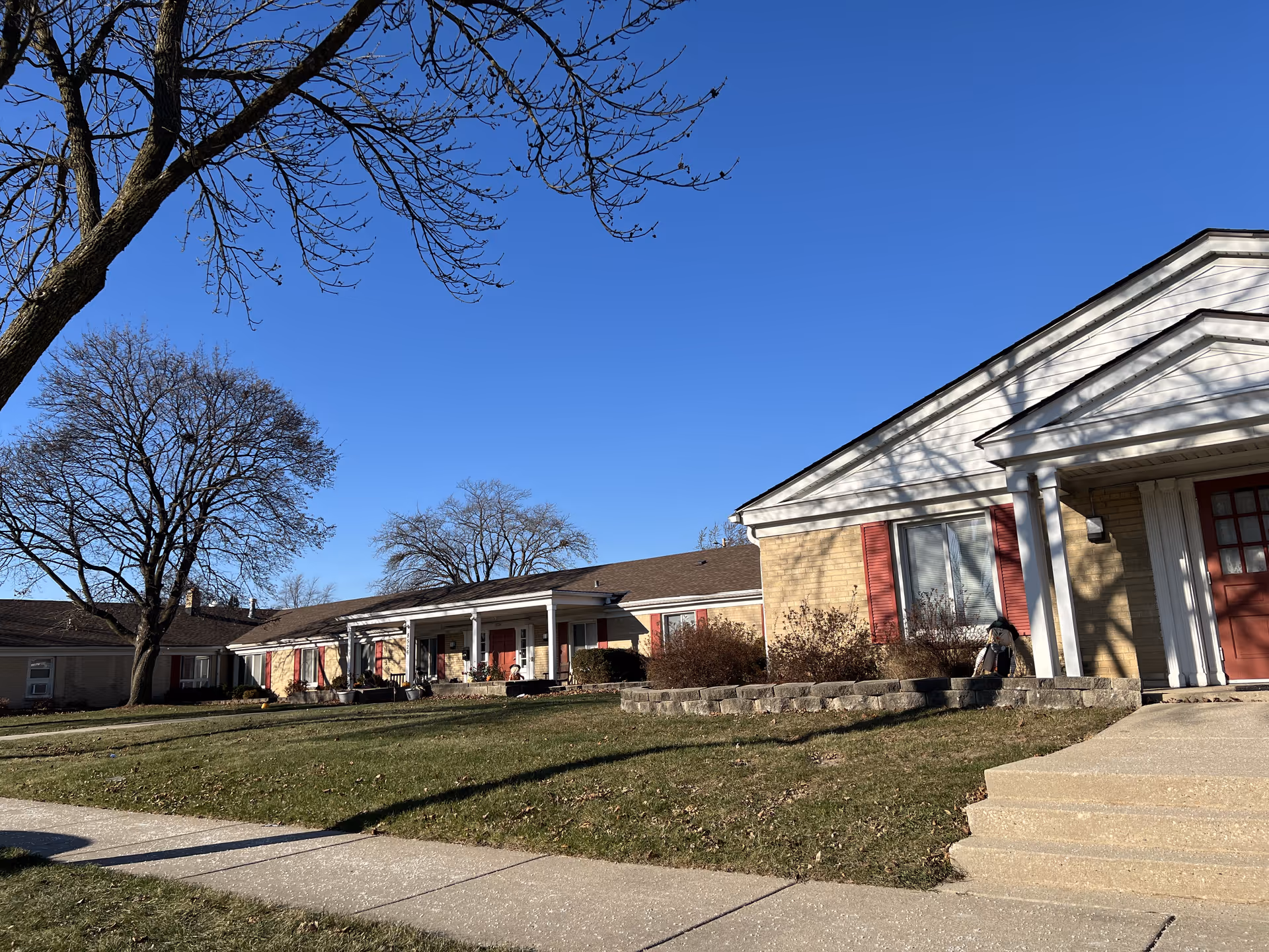 Exterior view of a single-story senior living facility building with beige brick walls, white trim, and red shutters. There is a small porch area with white columns and a red door. Leafless trees and a clear blue sky are visible in the background, along with a grassy lawn and a concrete sidewalk in the foreground.