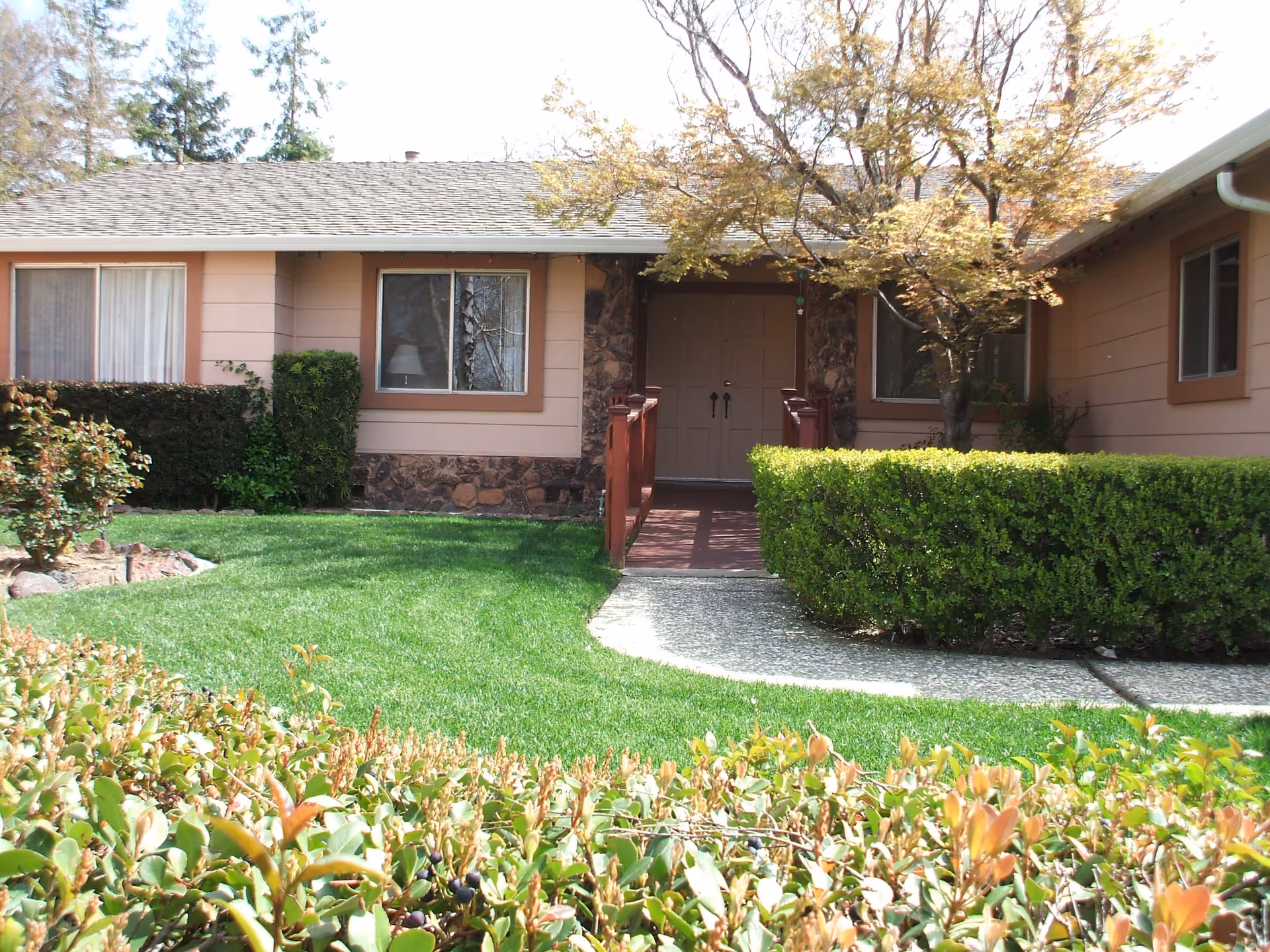 Front exterior view of a single-story building with beige walls, brown trim, and a shingled roof. There is a pathway leading to double front doors, surrounded by well-maintained green bushes, a tree with light brown leaves, and a green lawn.