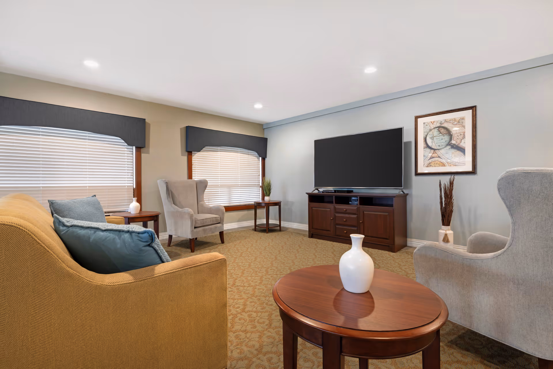 A cozy living room with a beige sofa adorned with blue pillows, two gray armchairs, a wooden coffee table with a white vase, a wooden TV stand with a large flat-screen television, two windows with blinds and dark valances, a framed map on the wall, and a decorative plant in a vase on the floor.